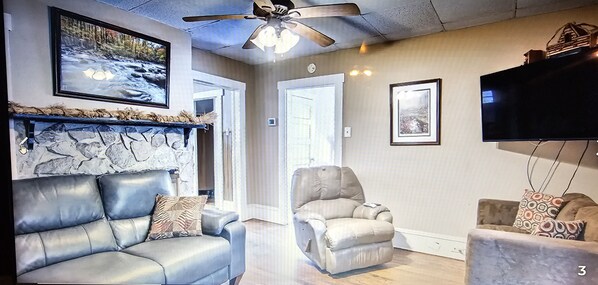 A living room with a stone fireplace, a ceiling fan, and a television mounted on the wall.