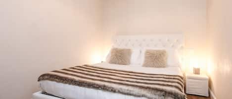 Una cama bien tendida con una colcha a rayas y dos almohadas, un cabecero blanco y una mesita de noche con una lámpara.