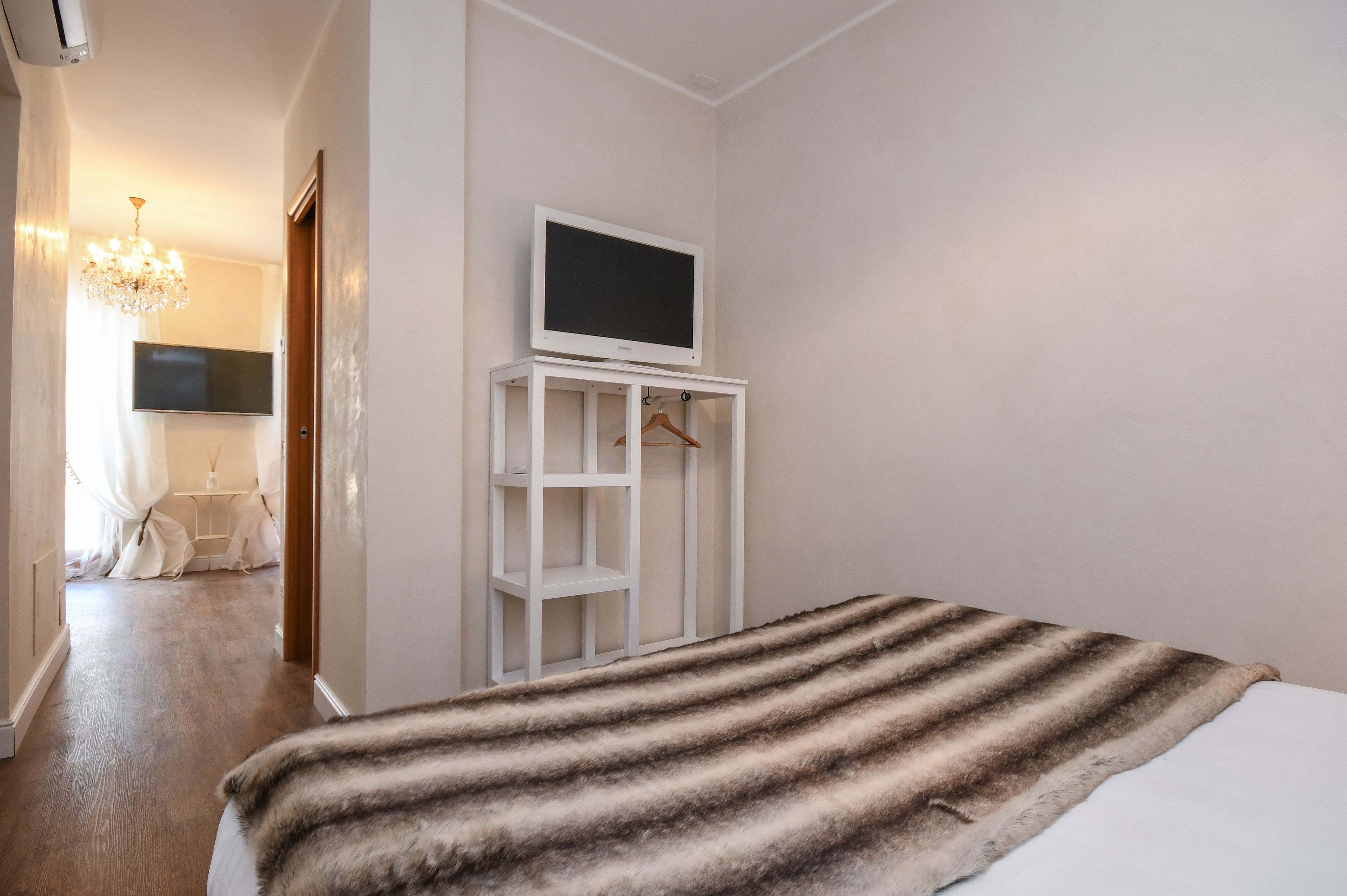 A bedroom with a bed, a TV on a stand, a chandelier, and a hallway leading to another room.