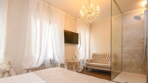 Un dormitorio con una cama grande, una mesita de noche, un televisor, una araña y un sofá, además de un vestidor con ducha.