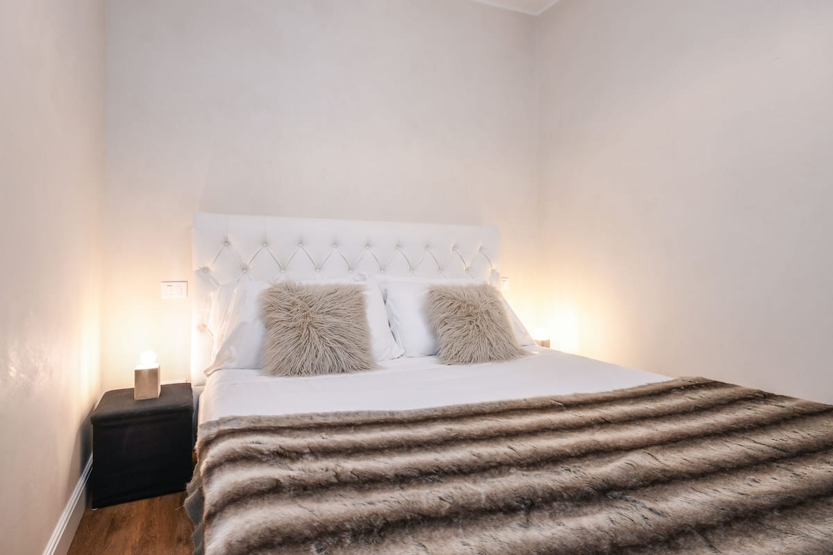 A neatly made bed with a tufted headboard, two fluffy pillows, a dark nightstand with a lamp, and a fur throw on the bed.