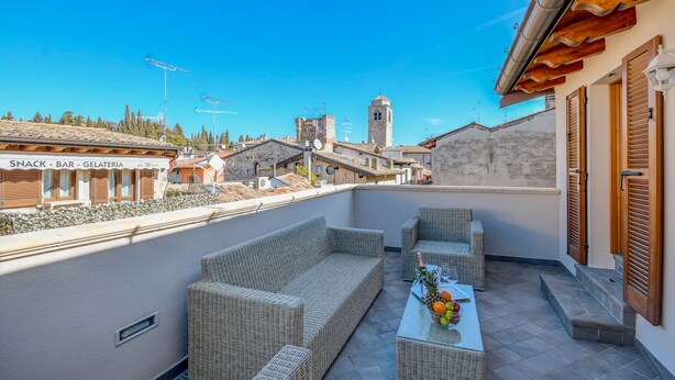 Terraza en la azotea con mobiliario de mimbre, una mesa con fruta y vistas al pueblo, incluyendo una torre y edificios.