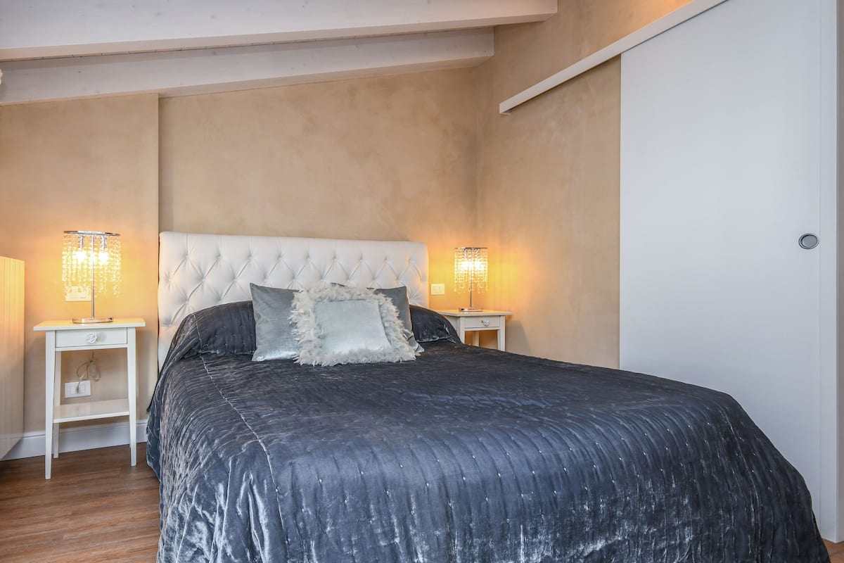 A bedroom with a tufted headboard, a dark bedspread, two bedside lamps, and a white nightstand.