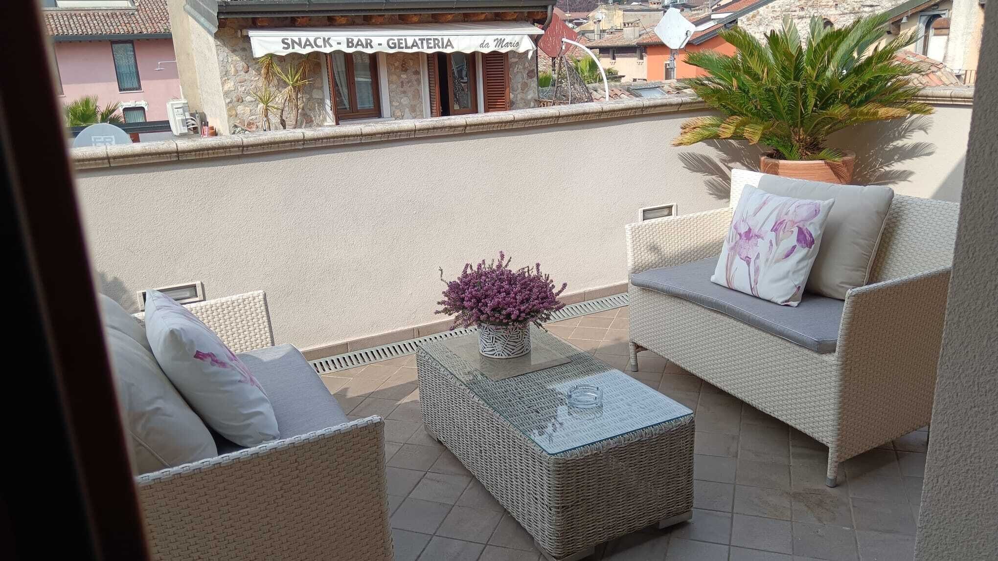 Un espace extérieur aménagé avec des meubles en osier, un pot de fleurs et une vue sur un snack-bar situé de l’autre côté de la rue.