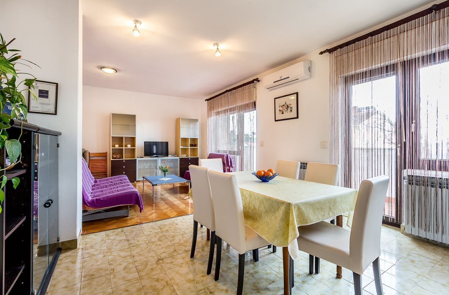 A hotel room with a dining area, a TV, a sofa, and a balcony with curtains.