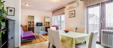 A hotel room with a dining area, a TV, a sofa, and a balcony with curtains.