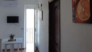 A hallway with a white door, a painting of a citrus fruit, a mounted TV, and a potted plant on a table.