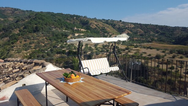 A wooden table with a basket of fruit and a bench with a striped cushion on a balcony overlooking a hilly landscape.