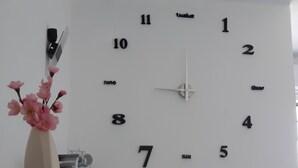 A wall clock with numbers and words indicating time, a vase with pink flowers, and a kitchen area with a coffee machine.