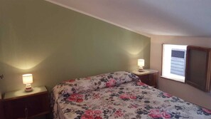 Une chambre à coucher avec une couvre-lit à motifs floraux, deux lampes de chevet, une fenêtre avec des stores et une tête de lit en bois.