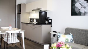 A modern kitchen with white cabinets, a black countertop, and a stainless steel refrigerator. There is a dining area with a white table and chairs, and a gray sofa with a colorful pillow. - Appartement Neuf et équipé, près des Antilles (Jonzac)