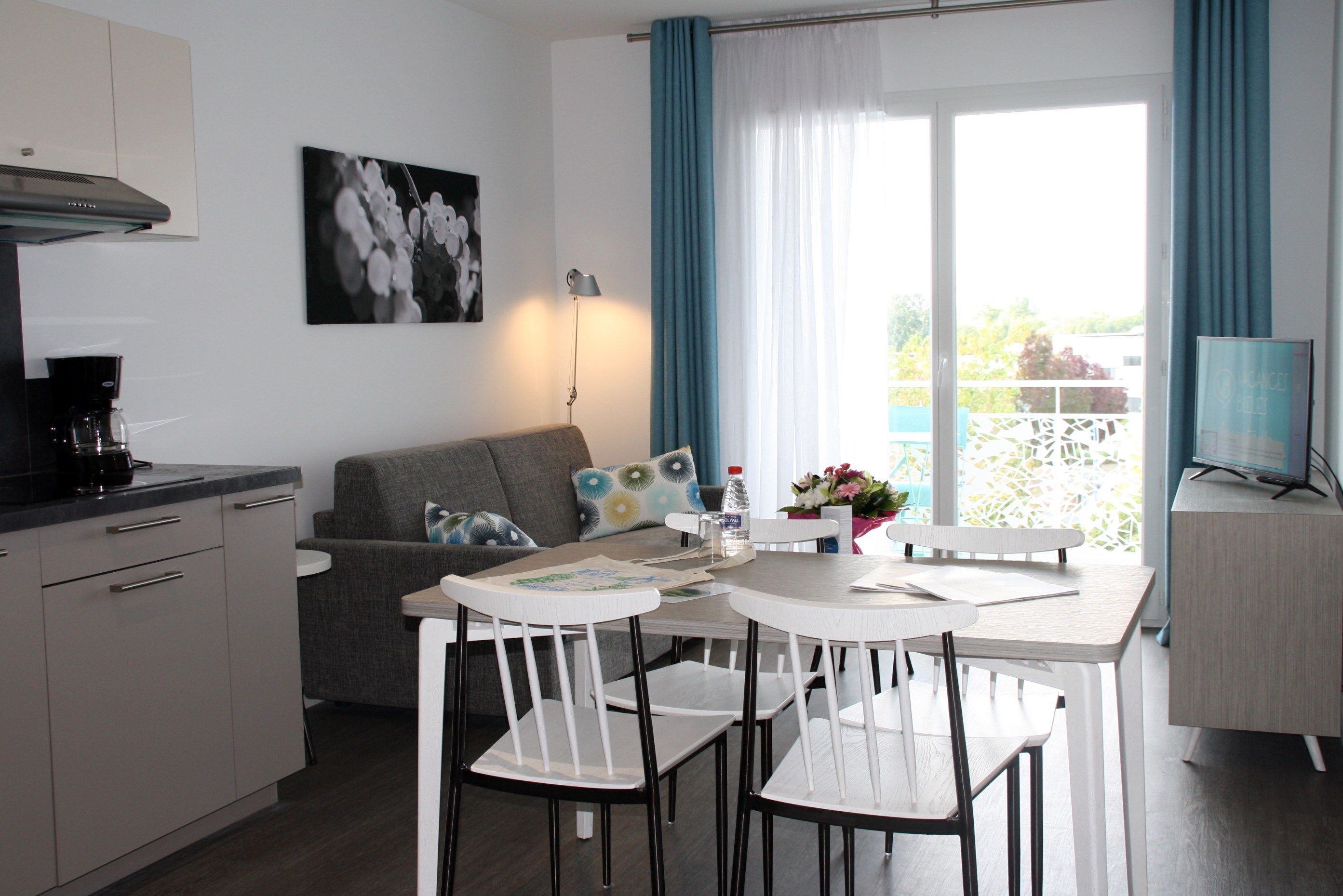 A modern kitchen with a coffee maker, a dining area with white chairs and a table, a grey sofa with decorative pillows, and a balcony with blue curtains.