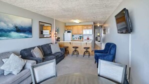 Un espacio habitable compacto con sofá, una mesa de comedor, un televisor montado en la pared y una zona de cocina con armarios de madera y un microondas.