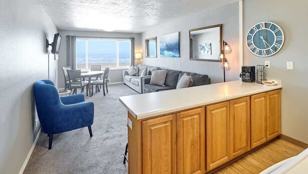 Una sala de estar moderna con un ventanal grande, un sillón azul, un sofá, una zona de comedor y una encimera de cocina.
