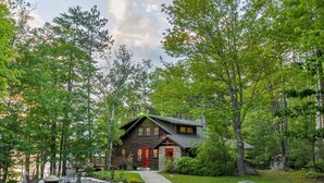 A cabin with a stone foundation, a wooden exterior, and a red door. There is a paved driveway leading to the entrance, surrounded by trees and a small garden area.