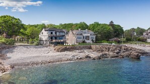 A large, multi-story house with a balcony, situated by a rocky shoreline with clear water.