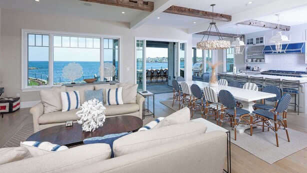 A spacious living area with a view of the sea, a dining area with blue and white patterned chairs, and a modern kitchen with white cabinets.