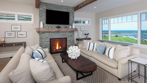 A living room with a stone fireplace, a large TV, and a view of the ocean.