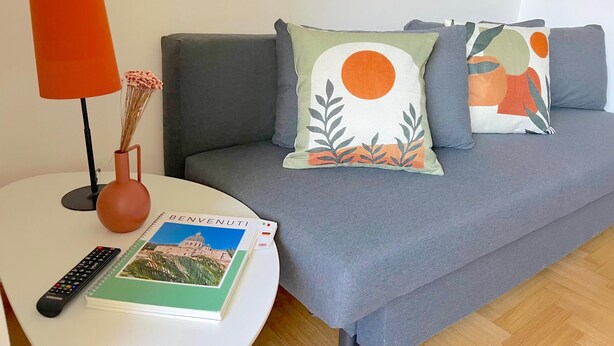 A grey sofa with decorative pillows, a white side table with a book and remote control, and a red lamp. - Colourful apartment  St. Peter's View (ROMA)