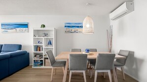En moderne stue med en blå sofa, et træspisebord med grå stole, en hvid reol med diverse genstande og to billeder på væggen.