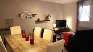 A dining area with a wooden table, four chairs, a TV, and a shelf with flowers.