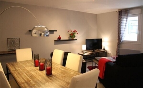 Uma sala de jantar com uma mesa de madeira, quatro cadeiras, uma televisão e uma estante com flores.