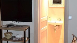 Una habitación con una televisión sobre un mueble bajo, una estantería con diversos objetos, y una puerta abierta que da a un baño con lavamanos y espejo.