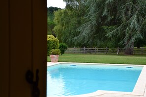 A swimming pool with a wooden deck and a view of a grassy area with trees and a fence. - Domaine Le Mathi (Fumel)