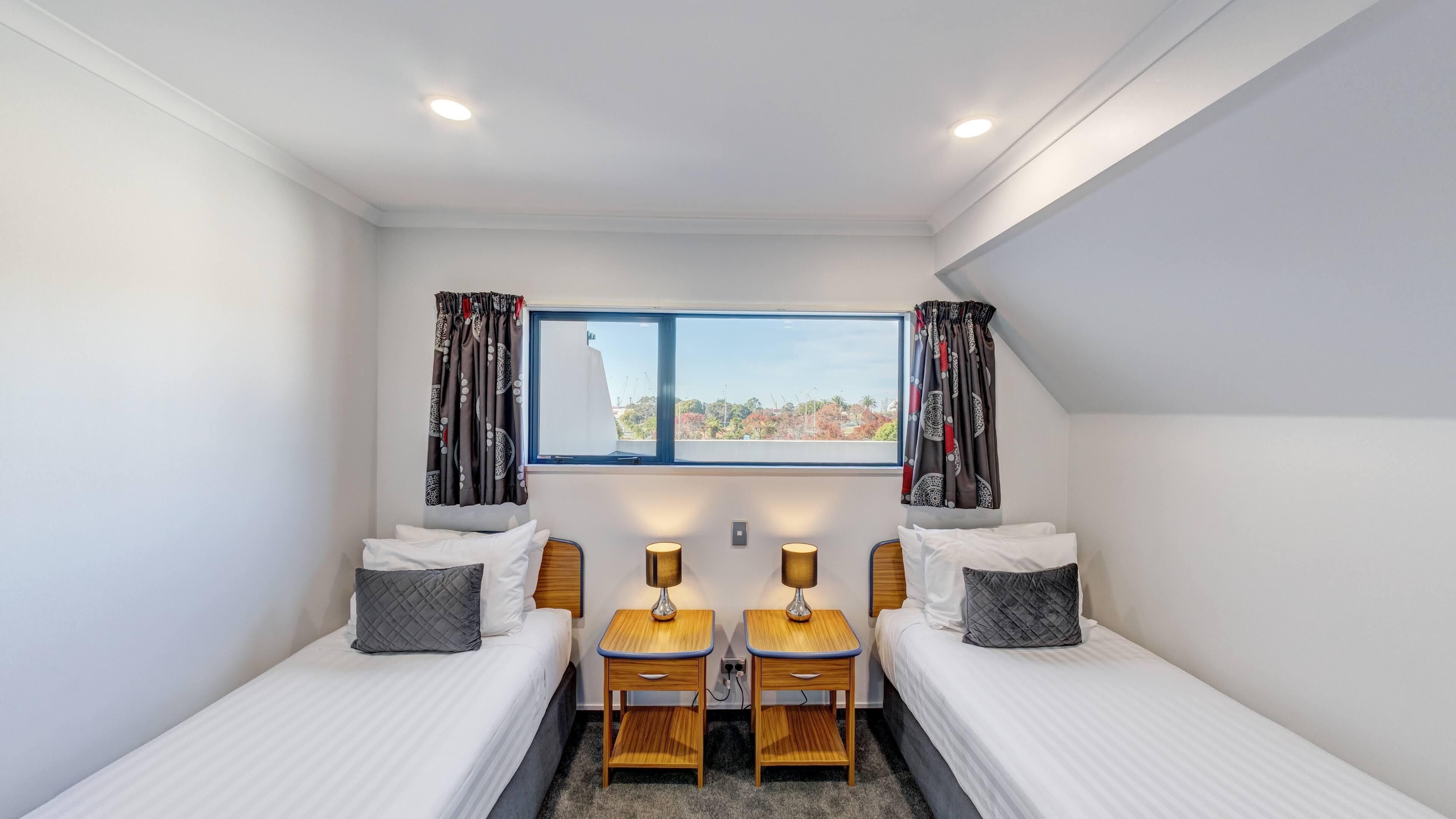 A small, well-lit bedroom with two single beds, a window with curtains, and two bedside tables with lamps.
