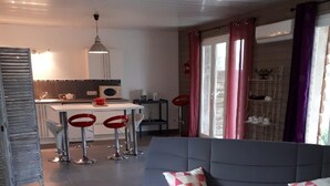A modern living room with a grey sofa, a black coffee table, and a red and white patterned pillow. There is a kitchen area in the background with a white countertop and red bar stools.