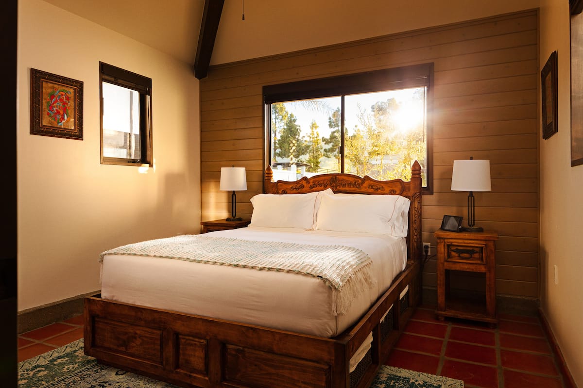 Una cama de madera con colchón y almohadas blancas, flanqueada por dos mesitas de noche con lámparas, en una habitación con paredes de madera y una ventana con vistas a árboles.