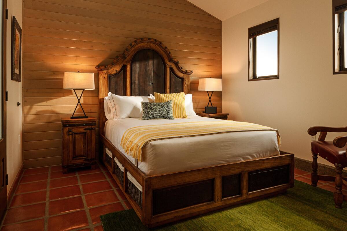 Una cama con estructura de madera y cabecera, dos mesitas de noche con lámparas, una silla y un ventanal.