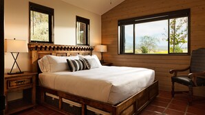 A wooden-framed bed with a white mattress, two bedside tables with lamps, a chair, and a window with a view of trees and mountains. - La Lomita Ranch (San Luis Obispo)