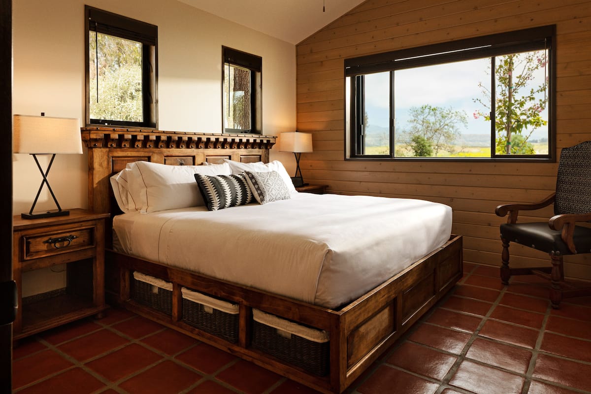 Una cama con estructura de madera y colchón blanco, dos mesitas de noche con lámparas, una silla y una ventana con vistas a árboles y montañas.