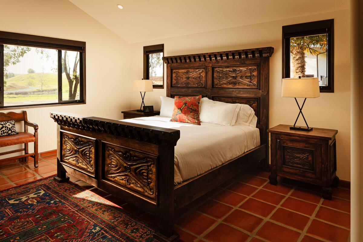 Un dormitorio con una cama grande, dos mesitas de noche, una silla y vistas al exterior.