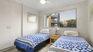 Une chambre avec deux lits, une fenêtre donnant sur un balcon, une table de chevet avec une lampe et un tableau accroché au mur.