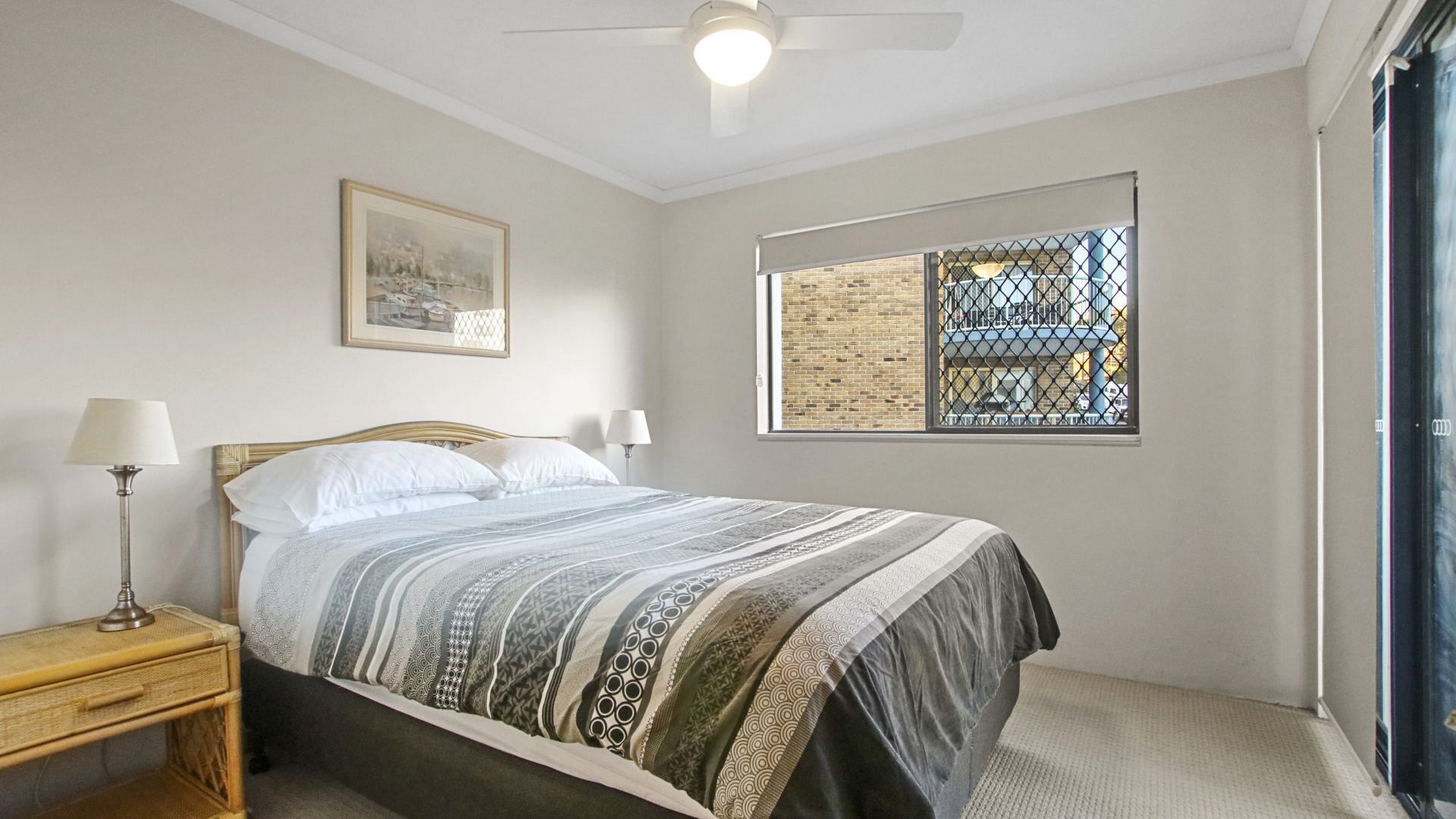 A bedroom with a bed, a nightstand, a lamp, a ceiling fan, a window with a view of a brick building, and a framed picture on the wall.
