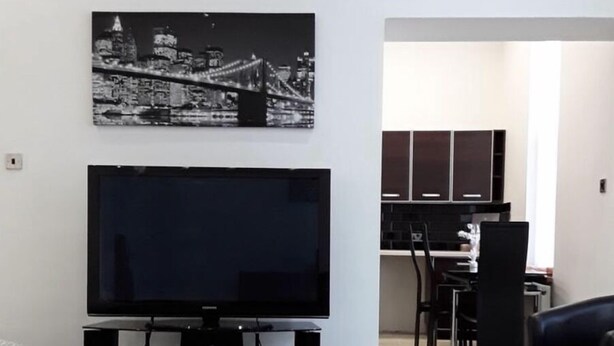 Una sala de estar con un televisor de pantalla plana sobre un mueble bajo, un comedor con mesa y sillas, y una cocina con armarios.