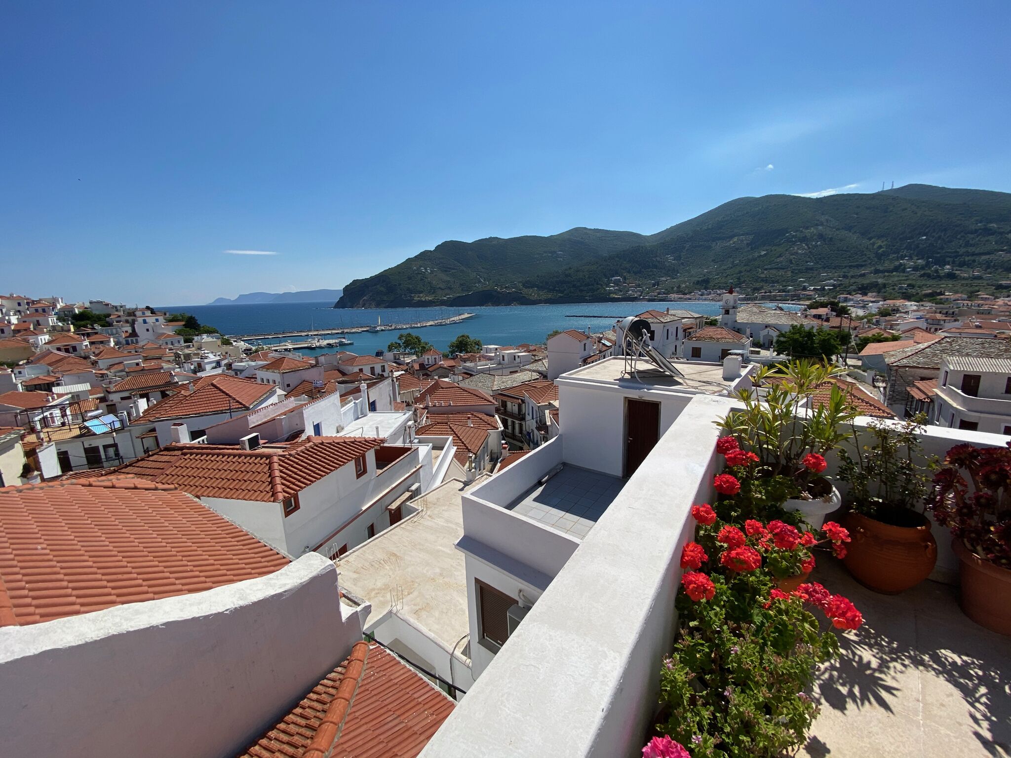 A coastal town with terracotta-roofed buildings, a marina, and a mountainous backdrop.