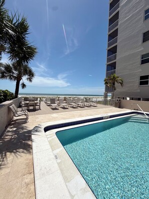 A clear blue pool with lounge chairs and tables, a palm tree, and a view of the ocean.