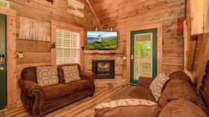 A cozy wooden cabin interior with a fireplace, two leather sofas, and a television displaying a scenic view. - 🐾A Mountain Heart Song🔥Pet-Friendly, Fire Pit, Hot Tub & Gazebo (Sevierville)