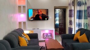A living room with a flat-screen TV mounted on the wall, two sofas with cushions, a wooden coffee table, and a ceiling fan. - Sunset Paradise Baringo 12 (Mombasa)