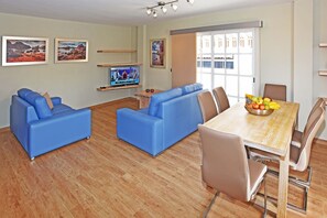 Un salon moderne avec des canapés bleus, une table à manger en bois sur laquelle on a disposé des fruits, et un téléviseur posé sur un meuble.