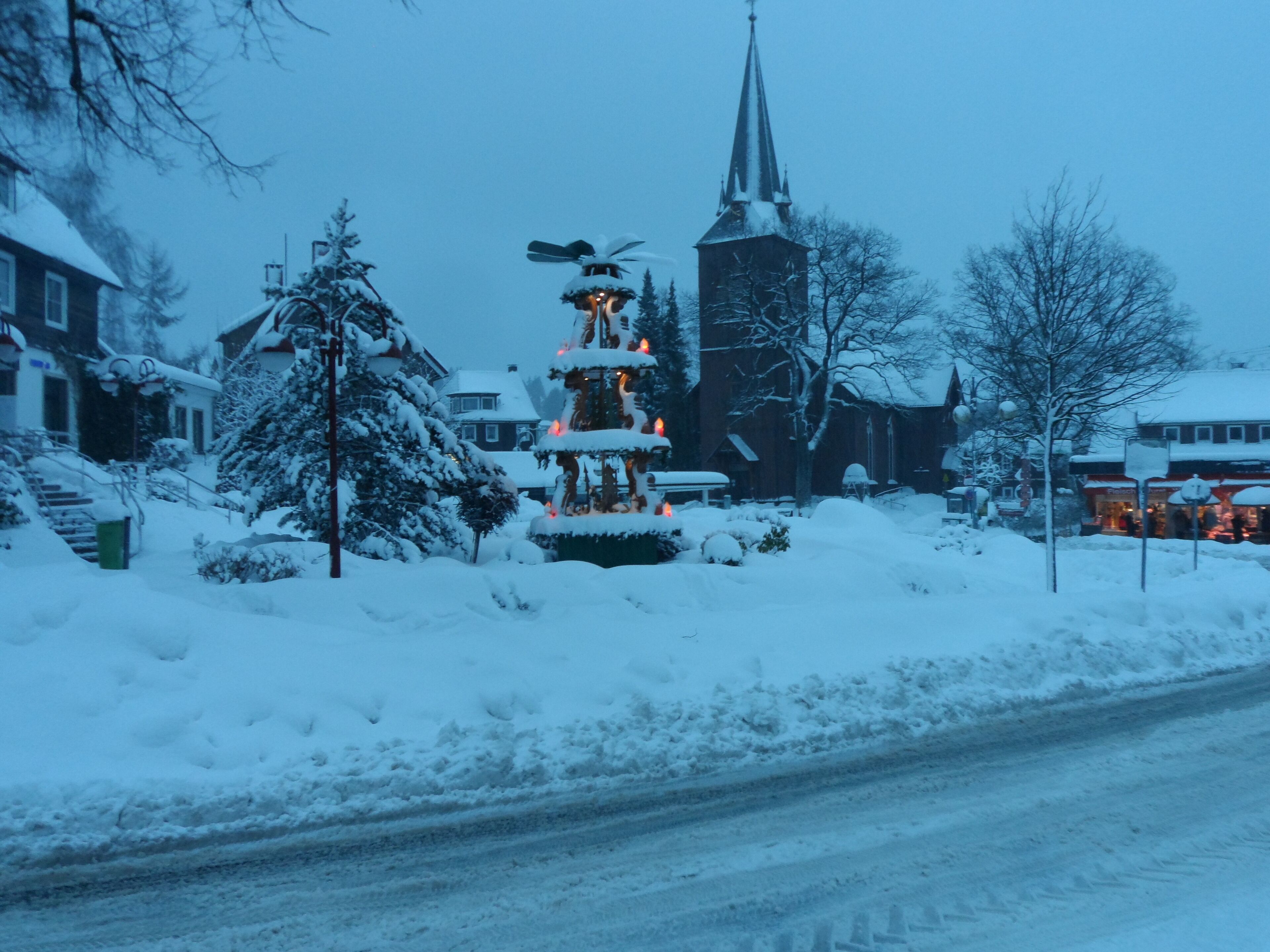 Una plaza nevada con una aguja de iglesia, un árbol de Navidad y una fuente.