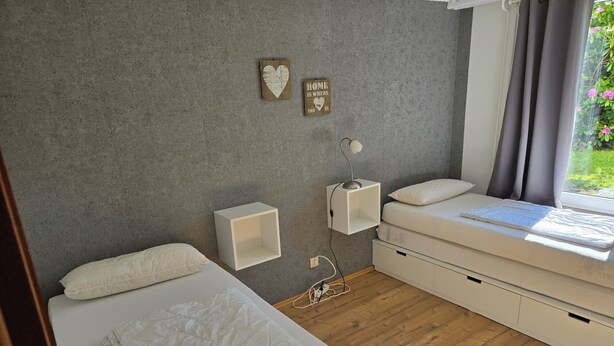 A bedroom with a bed, bedside tables, a wall-mounted shelf, a window with curtains, and a small plant.