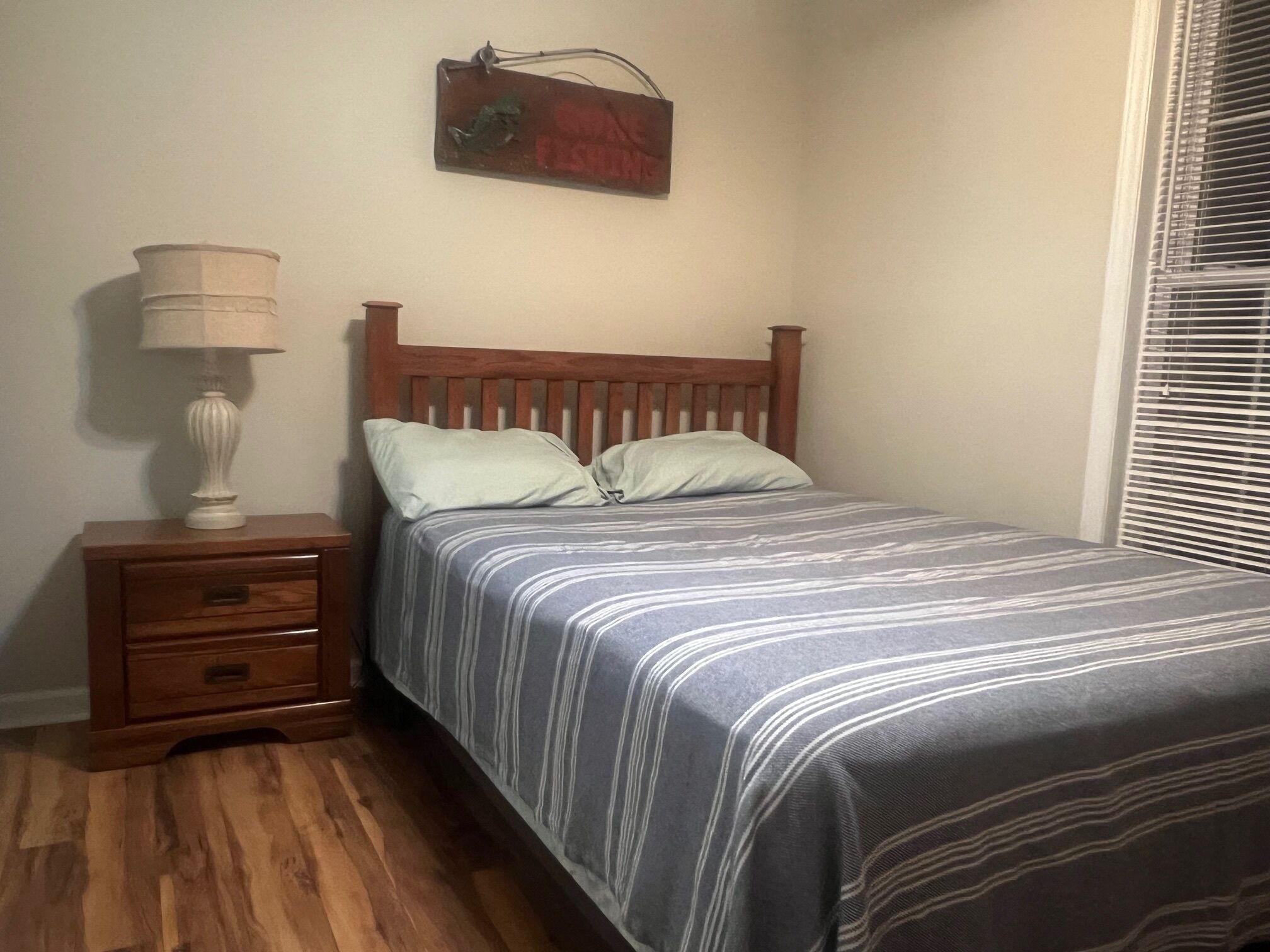 Een slaapkamer met een houten bed, twee kussens, een nachtkastje met een lamp en een aan de muur hangend schilderij.