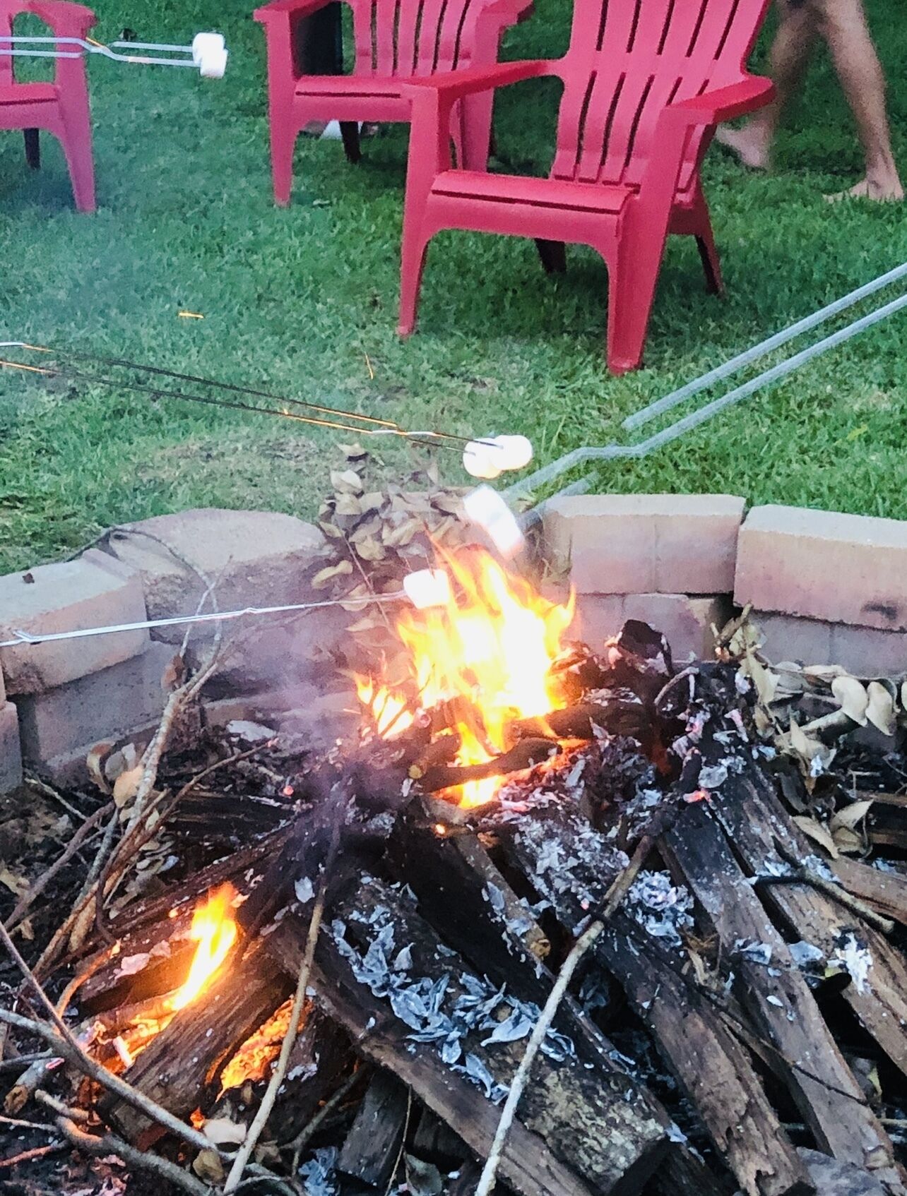 A campfire with marshmallows roasting, surrounded by a brick fire pit and red plastic chairs in a grassy area.