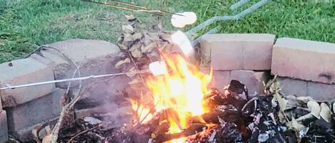 A campfire with marshmallows roasting, surrounded by a brick fire pit and red plastic chairs in a grassy area.