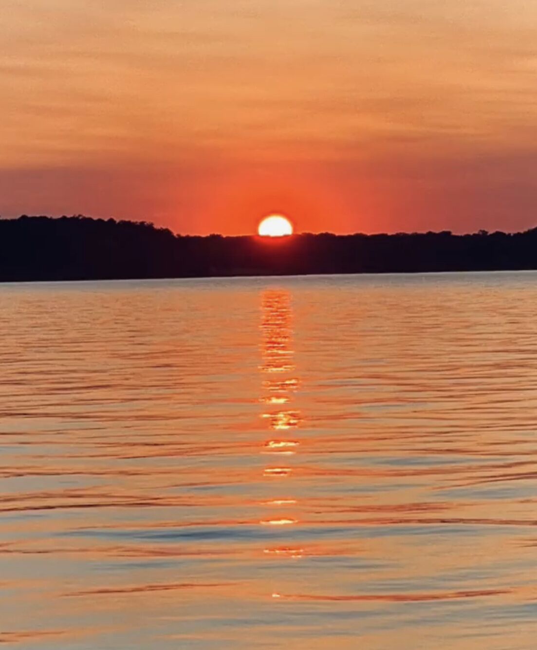 A sunset over a calm body of water with a silhouette of trees in the background.