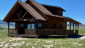 A wooden cabin with a large porch and a steeply pitched roof, surrounded by a grassy field and mountains. - Montana Prairie Cabin (McAllister)
